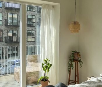 A sunlit bedroom with airy curtains and a mirror reflecting a lush indoor plant.